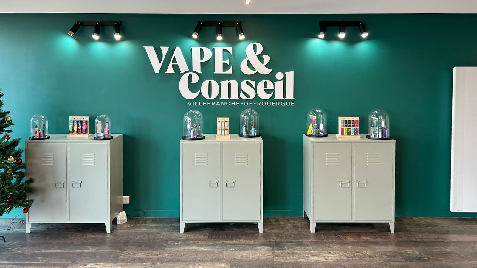 Le mur vert de la boutique avec l’enseigne Vape & Conseil et trois meubles présentant du matériel sous cloches de verre.