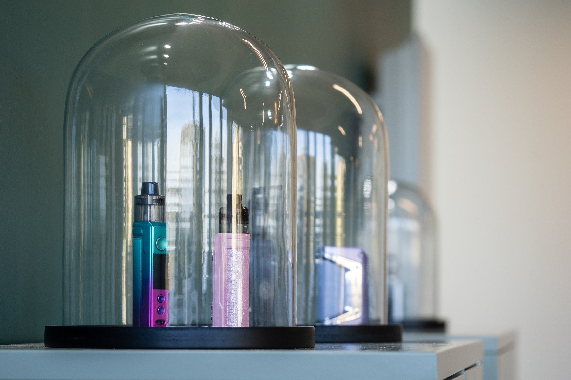 Deux appareils de vapotage présentés sous des cloches de verre, sur un meuble clair.